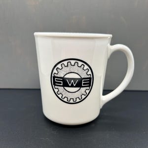 May include: White ceramic mug with a black and white gear design and the letters "SWE". The mug has a curved handle and a slightly flared rim. The mug is on a dark surface.