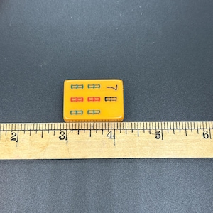 May include: A close-up of a yellow Mahjong tile with the number 7. The tile has three rows of symbols in green, red, and green. A wooden ruler is next to the tile, with measurements in inches and centimetres.
