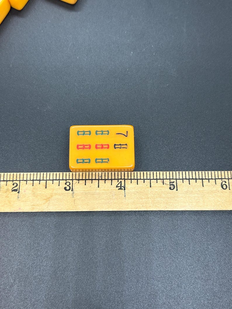 May include: A close-up of a yellow Mahjong tile with the number 7. The tile has three rows of symbols in green, red, and green. A wooden ruler is next to the tile, with measurements in inches and centimetres.