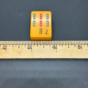 May include: A vintage, rectangular Mahjong tile in a yellow-orange colour. The tile displays a pattern of green and red symbols, with the number 7 in yellow. The tile is next to a wooden ruler.