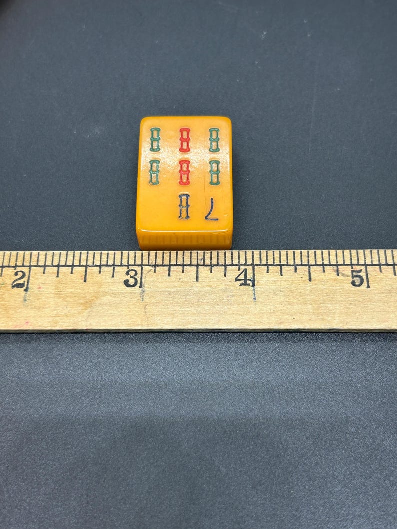 May include: A vintage, rectangular Mahjong tile in a yellow-orange colour. The tile displays a pattern of green and red symbols, with the number 7 in yellow. The tile is next to a wooden ruler.