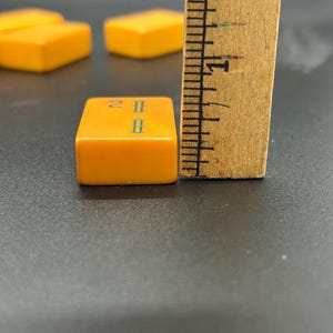 May include: Close-up of a vintage orange Mahjong tile with green markings, measuring approximately 1 inch. The tile is next to a wooden ruler. Other tiles are visible in the background.