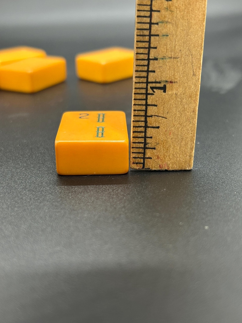 May include: Close-up of a vintage orange Mahjong tile with green markings, measuring approximately 1 inch. The tile is next to a wooden ruler. Other tiles are visible in the background.