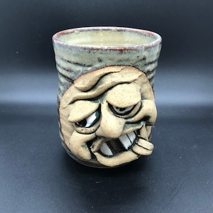 May include: Handmade ceramic mug with a sculpted face design. The mug features a textured, cylindrical body with a light green and brown glaze. The face has exaggerated features, including a wide grin and bulging eyes, adding a unique, artistic touch to the drinkware.
