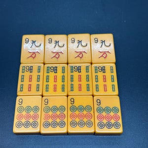 Op de afbeelding: Een verzameling vintage Mahjong-tegels. De tegels zijn lichtgeel met zwarte, rode, groene en blauwe symbolen. De tegels zijn gerangschikt in drie rijen van vier.