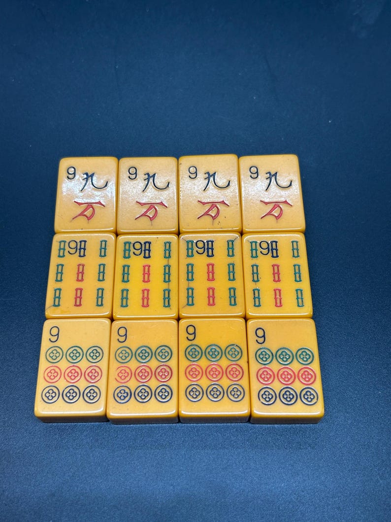 Op de afbeelding: Een verzameling vintage Mahjong-tegels. De tegels zijn lichtgeel met zwarte, rode, groene en blauwe symbolen. De tegels zijn gerangschikt in drie rijen van vier.