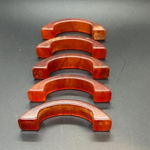 May include: Five semi-circular, arch-shaped objects made of a translucent, deep orange material with marbled, amber-like patterns. Each piece features a smooth, curved top edge and flat, squared-off ends, arranged in a vertical row on a dark surface.