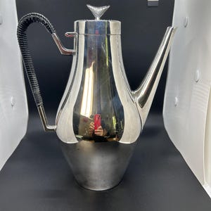 May include: A tall, polished silver-tone metal coffee pot with a tapered body, a long curved spout, and a lid topped with a small finial. The handle is wrapped in black textured material, providing a contrast to the reflective metallic surface.