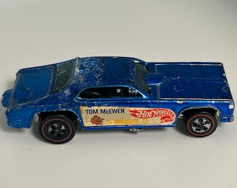 May include: Blue diecast toy car with red wheels and "Hot Wheels" and "The Mongoose II" printed on the side. The car is a replica of a 1970s muscle car.
