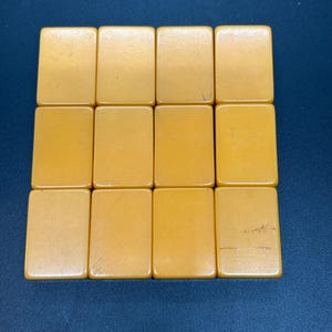 Op de afbeelding: Een verzameling van twaalf rechthoekige, amberkleurige tegels, gerangschikt in drie rijen van vier. De tegels hebben een gladde, glanzende afwerking en lijken van een massief materiaal te zijn gemaakt. De tegels staan tegen een donkerblauwe achtergrond.