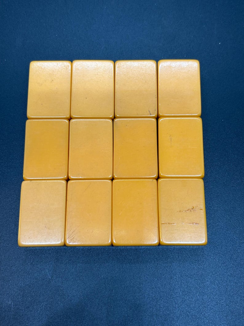 Op de afbeelding: Een verzameling van twaalf rechthoekige, amberkleurige tegels, gerangschikt in drie rijen van vier. De tegels hebben een gladde, glanzende afwerking en lijken van een massief materiaal te zijn gemaakt. De tegels staan tegen een donkerblauwe achtergrond.