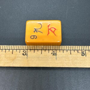 Op de afbeelding: Een oranje rechthoekige Mahjong-tegel met zwarte en rode symbolen. De tegel is ongeveer 3,8 cm lang en staat tegen een donkere achtergrond. Een houten liniaal is naast de tegel geplaatst.