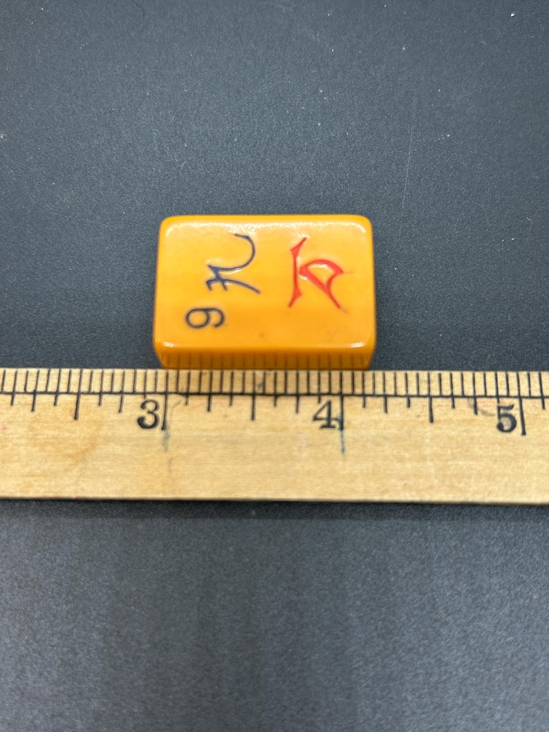 Op de afbeelding: Een oranje rechthoekige Mahjong-tegel met zwarte en rode symbolen. De tegel is ongeveer 3,8 cm lang en staat tegen een donkere achtergrond. Een houten liniaal is naast de tegel geplaatst.