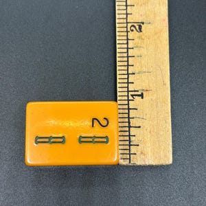 May include: An orange rectangular tile with the number 2 and two green horizontal lines. A wooden ruler is next to the tile, showing measurements in inches.