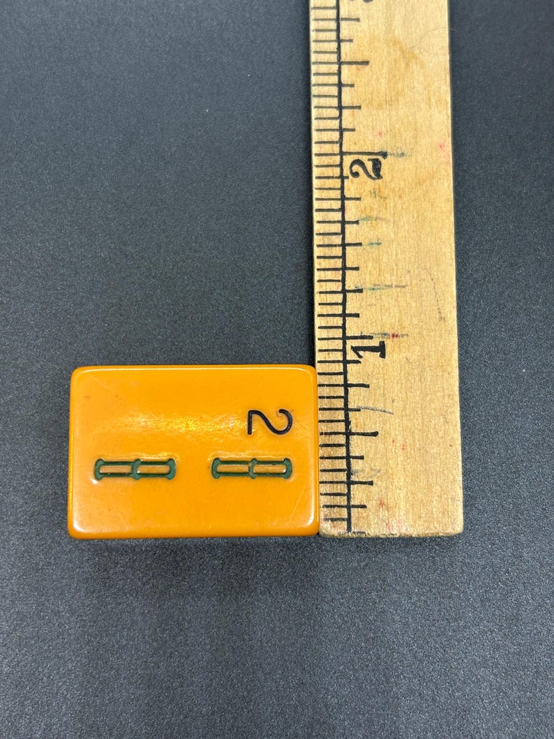 May include: An orange rectangular tile with the number 2 and two green horizontal lines. A wooden ruler is next to the tile, showing measurements in inches.