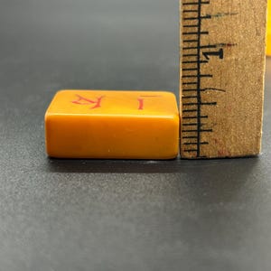 May include: Close-up of a rectangular, orange Mahjong tile with red Chinese characters. A wooden ruler is positioned next to the tile, showing inch measurements. The tile is on a dark surface.