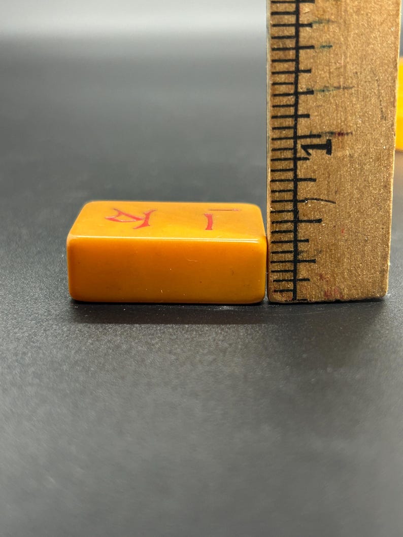 May include: Close-up of a rectangular, orange Mahjong tile with red Chinese characters. A wooden ruler is positioned next to the tile, showing inch measurements. The tile is on a dark surface.