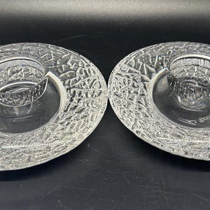 May include: Two clear glass candle holders with a textured, oval design. Each has a central, circular well for a candle. The glass has a textured pattern. The candle holders are on a dark surface.