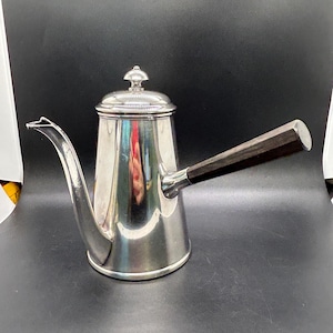 May include: A silver-colored coffee pot with a dark wooden handle. The pot has a long, curved spout and a lid with a small knob. The pot is reflective, showing the surrounding environment.