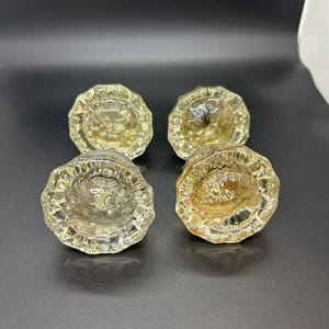 May include: A set of four clear glass door knobs, each featuring a multi-faceted, circular design with a scalloped edge. The knobs are transparent with a reflective quality, showcasing a starburst pattern at the center of each piece.