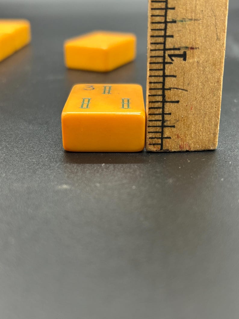 May include: Close-up of a vintage orange Mahjong tile with the number 3 and two 8s in blue. A wooden ruler is next to the tile. Other orange tiles are in the background.