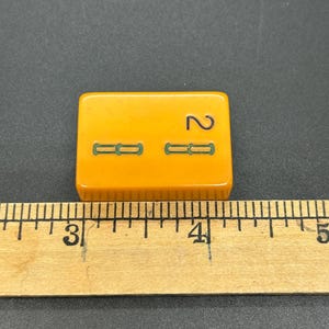 May include: A vintage, rectangular, orange tile with the number 2 and two sets of green dashes. The tile is approximately 1.5 inches long. The tile is placed on a black surface next to a wooden ruler.