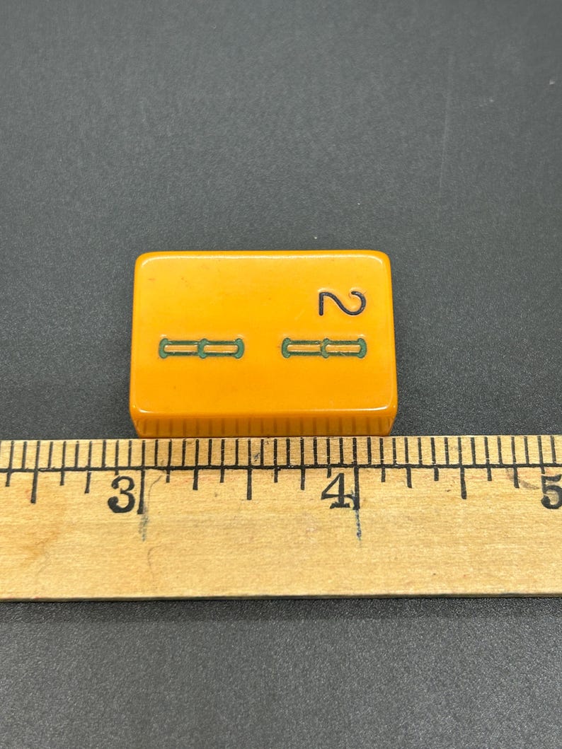 May include: A vintage, rectangular, orange tile with the number 2 and two sets of green dashes. The tile is approximately 1.5 inches long. The tile is placed on a black surface next to a wooden ruler.