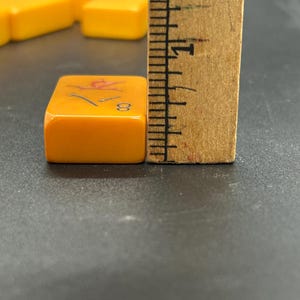 K&ouml;nnte beinhalten: Nahaufnahme einer orangefarbenen Mahjong-Kachel mit der Zahl 8 und einem roten Symbol. Die Kachel ist etwa 2 cm hoch und liegt neben einem Holzlineal. Weitere Kacheln sind im Hintergrund.