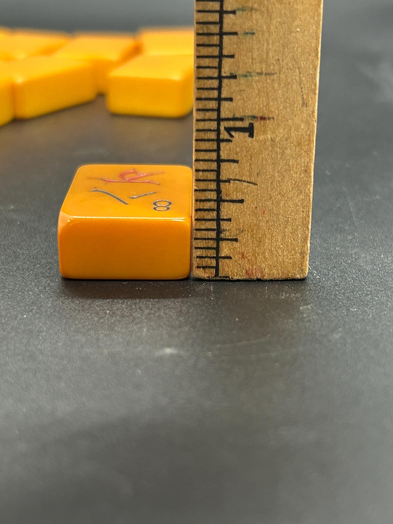 K&ouml;nnte beinhalten: Nahaufnahme einer orangefarbenen Mahjong-Kachel mit der Zahl 8 und einem roten Symbol. Die Kachel ist etwa 2 cm hoch und liegt neben einem Holzlineal. Weitere Kacheln sind im Hintergrund.
