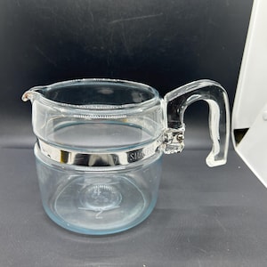 May include: A clear glass coffee pot with a stainless steel band and handle. The pot has a spout for pouring and a capacity marking of "2" on the bottom. The handle is clear and curved.