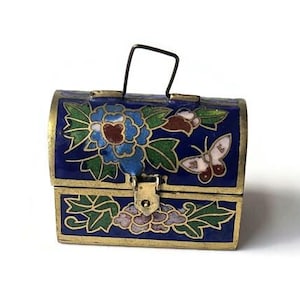 Puede incluir: Un pequeño cofre azul ornamentado con tapa con bisagras y asa negra. El cofre está decorado con diseños florales y de mariposas en tonos azules, verdes y dorados. El borde metálico es dorado.