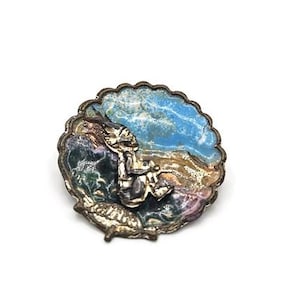 Könnte beinhalten: Vintage Brosche in Silberoptik mit gewelltem Rand. Die Brosche hat ein farbenfrohes Emaille-Design mit Blau-, Grün- und Brauntönen sowie eine kleine Figur. Ein einzigartiges Schmuckstück.