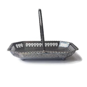 May include: An antique silver-toned metal basket with a handle. The basket has an openwork design with geometric patterns and sits on small feet. The handle is upright, and the basket is empty.