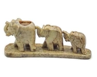May include: A carved stone sculpture of three elephants walking in a line. The elephants are light brown and beige, with a small hole in the back of the first elephant. The sculpture is on a rectangular base.