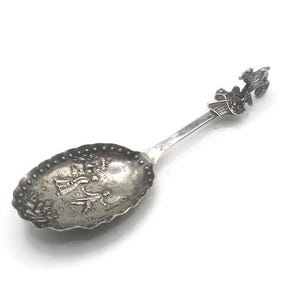May include: A silver-colored spoon with an ornate design. The spoon's bowl features a detailed embossed pattern, and the handle is topped with a decorative element. The spoon is likely a collectible or decorative item, possibly vintage.