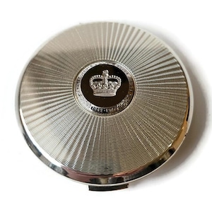 May include: A silver compact with a radial pattern. The centre has a black circle with a crown and the text "THE QUEEN'S SILVER JUBILEE 1952-1977". The compact has a hinged opening and a polished finish.