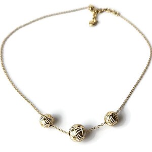 May include: A gold-colored necklace with three spherical, woven-style pendants. The delicate chain is adjustable and features a small heart-shaped charm near the clasp. The necklace is displayed against a white background.