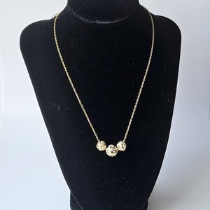 May include: A gold-colored necklace with three textured spherical pendants. The necklace is displayed on a black velvet bust. The pendants have a woven design, and the chain is delicate.