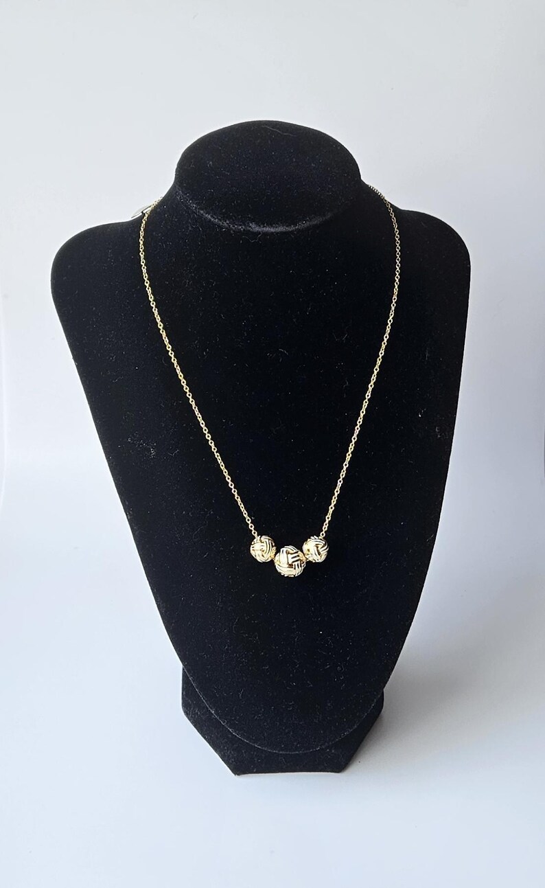 May include: A gold-colored necklace with three textured spherical pendants. The necklace is displayed on a black velvet bust. The pendants have a woven design, and the chain is delicate.