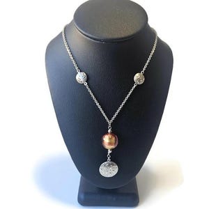 May include: Silver-toned necklace with a chain and three circular charms. The necklace features a copper-colored bead and a silver charm at the bottom. The charms have a decorative pattern. The necklace is displayed on a black jewelry stand.