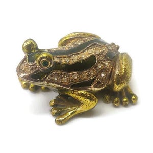 May include: A gold frog figurine with black and white stripes and rhinestones. The frog has green eyes and is sitting on a white surface.