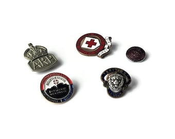 Op de afbeelding: Een verzameling vintage metalen spelden en badges. Bevat een rood kruis embleem, een British Legion badge met een leeuw en een badge met de letters "ARP" in een kroonontwerp. Andere badges bevatten tekst.