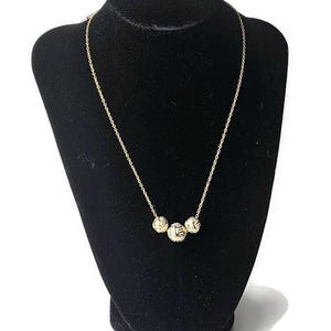 May include: A gold-colored necklace with three textured spherical pendants. The necklace is displayed on a black velvet bust against a white background. The chain is delicate and the pendants have a woven design.