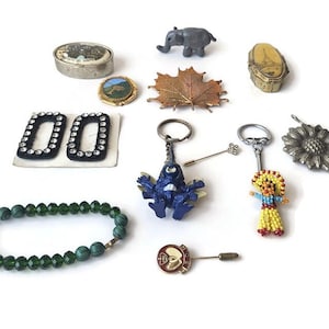 Puede incluir: Una colección de varias joyas y baratijas vintage, que incluyen una pulsera de cuentas, llaveros, broches y pequeñas cajas decorativas. Los artículos presentan diversos diseños, como una figura de elefante, un broche de hoja de arce y un llavero de muñeca de cuentas.