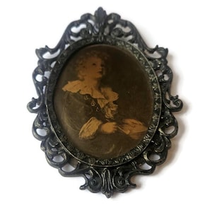May include: An antique oval portrait in a dark, ornate metal frame. The portrait features a sepia-toned image of a person with a ruffled collar. The frame has a detailed, scrollwork design, adding to its vintage aesthetic.