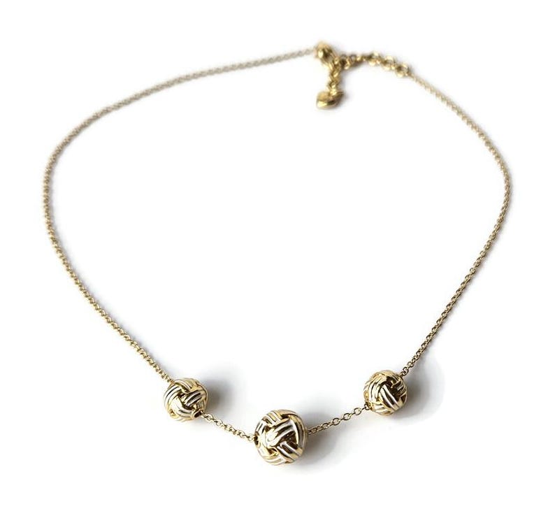 May include: A gold-colored necklace with three spherical, woven-style pendants. The delicate chain is adjustable and features a small heart-shaped charm near the clasp. The necklace is displayed against a white background.