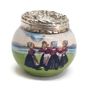 May include: A small, round ceramic jar with a silver-colored lid. The jar is painted with a scene of women in traditional clothing dancing in a circle. The background features a green field, blue water, and a light blue sky.