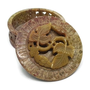 May include: A round, carved stone box with a lid. The lid features a design of two stylised leaves in a light brown colour. The box is made of a light brown stone with a mottled pattern.