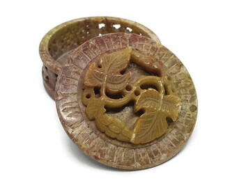May include: A round, carved stone box with a lid. The lid features a design of two stylized leaves in a light brown color. The box is made of a light brown stone with a mottled pattern.