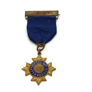 Pode incluir: Uma medalha dourada com um centro de esmalte azul que diz "Presença Regular" e um design de estrela. A medalha está presa a uma fita azul com um alfinete.